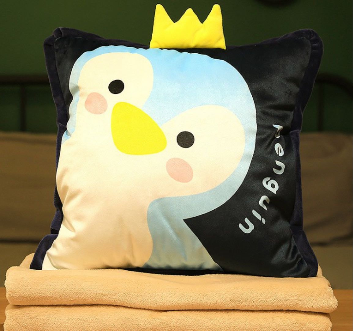 Close-up view of the blue and black plush penguin pillow featuring a yellow 3D crown on top and "Penguin" text embroidered on the side, stacked on beige blankets.