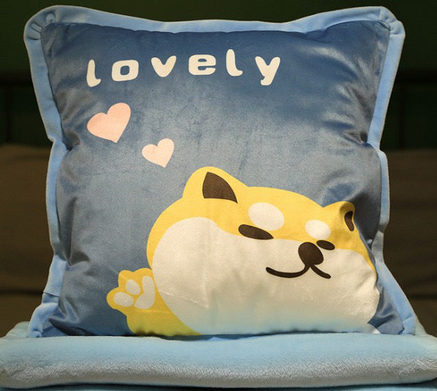 Blue plush pillow featuring a winking yellow Shiba Inu puppy, pink hearts, and "lovely" embroidered text, stacked on blue blankets.