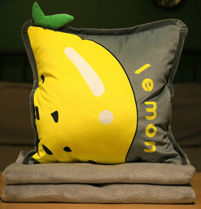 Grey and yellow plush pillow featuring a lemon graphic, "lemon" text, and a green 3D leaf on the corner, stacked on soft grey blankets.