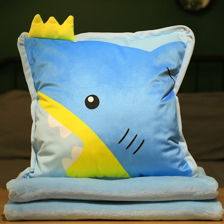 Blue plush pillow featuring a cartoon shark face with a yellow mouth and a 3D yellow crown, stacked on soft blue blankets.