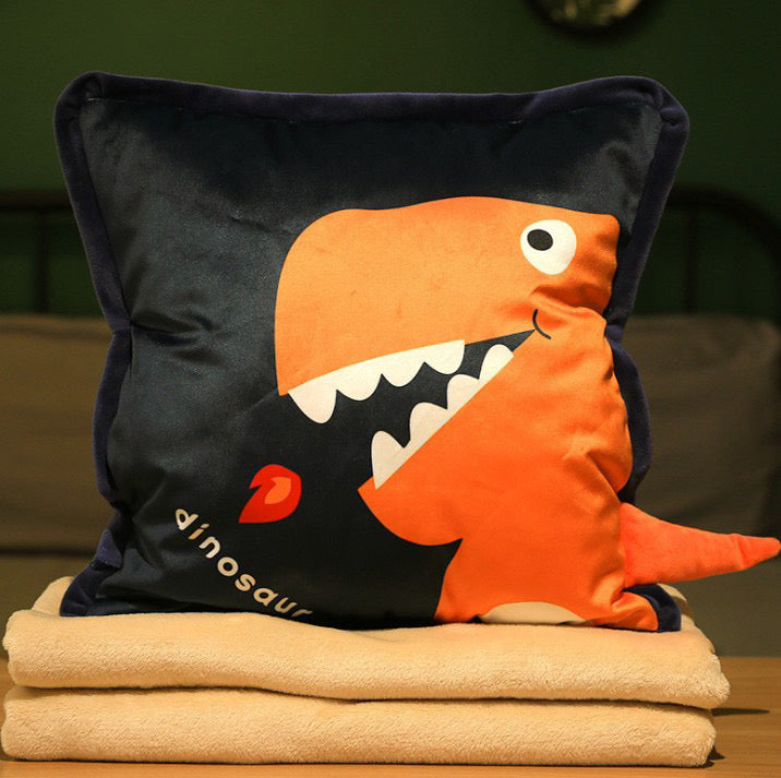 Navy blue plush dinosaur pillow featuring an orange T-Rex breathing fire with a 3D tail, stacked on beige blankets.