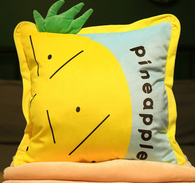 Bright yellow pineapple-shaped plush pillow with green 3D leaves and the word "pineapple" on the side, stacked on a soft blanke