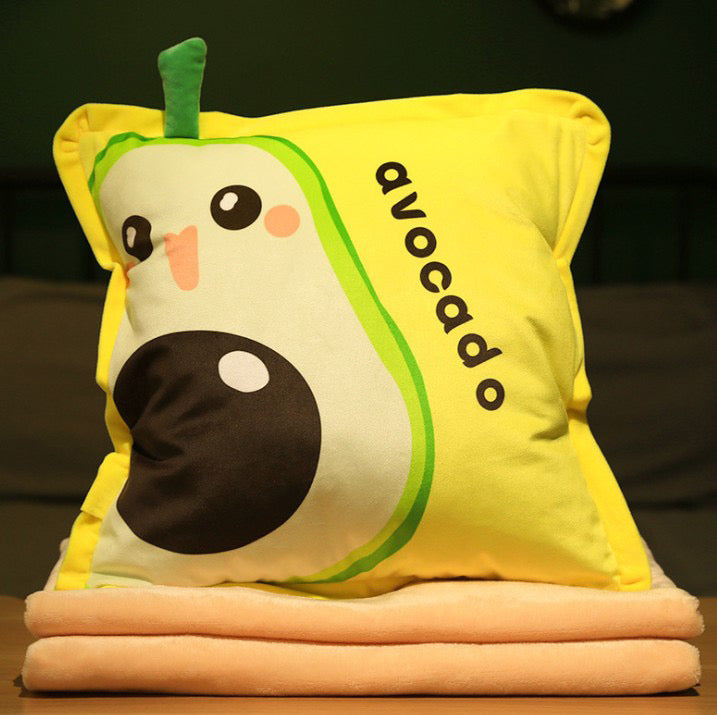 Bright yellow plush pillow featuring a cute cartoon avocado with a 3D green stem and "avocado" text, stacked on soft peach blankets.
